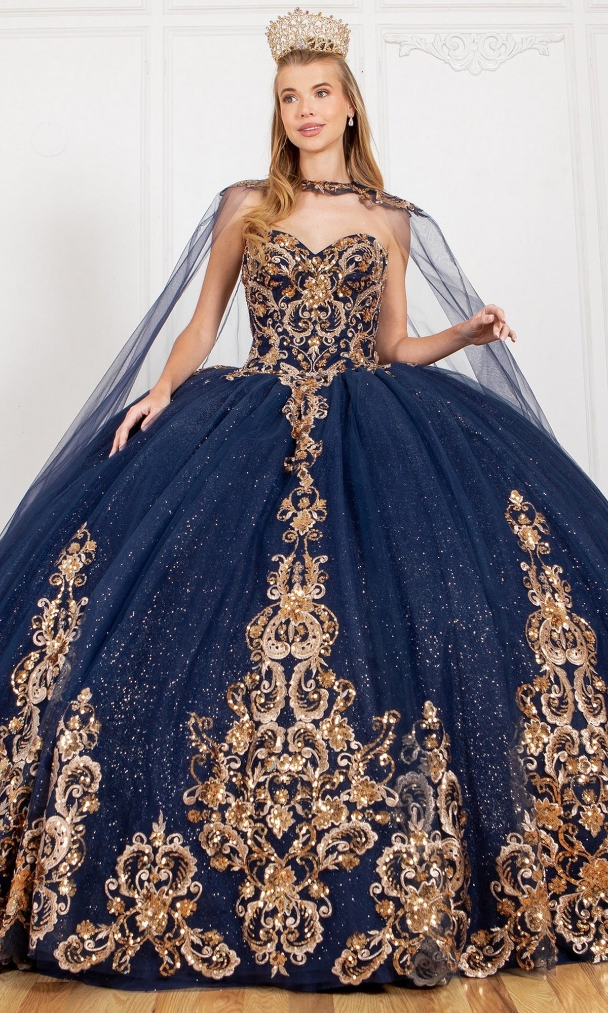 A woman in the Quinceanera Ball Gown 8063J, featuring dark blue fabric with gold embroidery, glitter tulle, and sparkle details, wears a jeweled gold crown and stands before a white ornate wall.